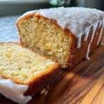Close-up of moist eggnog bread loaf with a thick white glaze dripping down the side, one slice cut.
