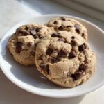 Three freshly baked peanut butter chocolate chip cookies stacked slightly on a white plate.
