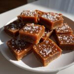 A pile of rich, brown salted caramel fudge squares topped generously with flaky sea salt.