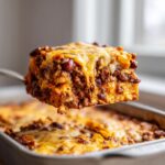 A spatula lifts a cheesy, layered slice of baked frito pie out of the pan.