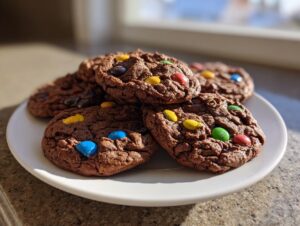 A stack of soft chocolate m&m cookies piled on a white plate, catching bright sunlight.