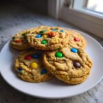 Four soft m&m cookies stacked slightly on a white plate near a window.
