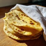 A warm stack of homemade corn tortillas, slightly browned, wrapped partially in a white cloth on a wooden surface.