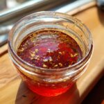 Close-up of a vibrant, reddish homemade salad dressing with visible herbs and garlic bits in a small glass jar.