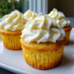 A close-up of three bright yellow lemon cupcakes topped with generous swirls of white frosting and lemon zest.