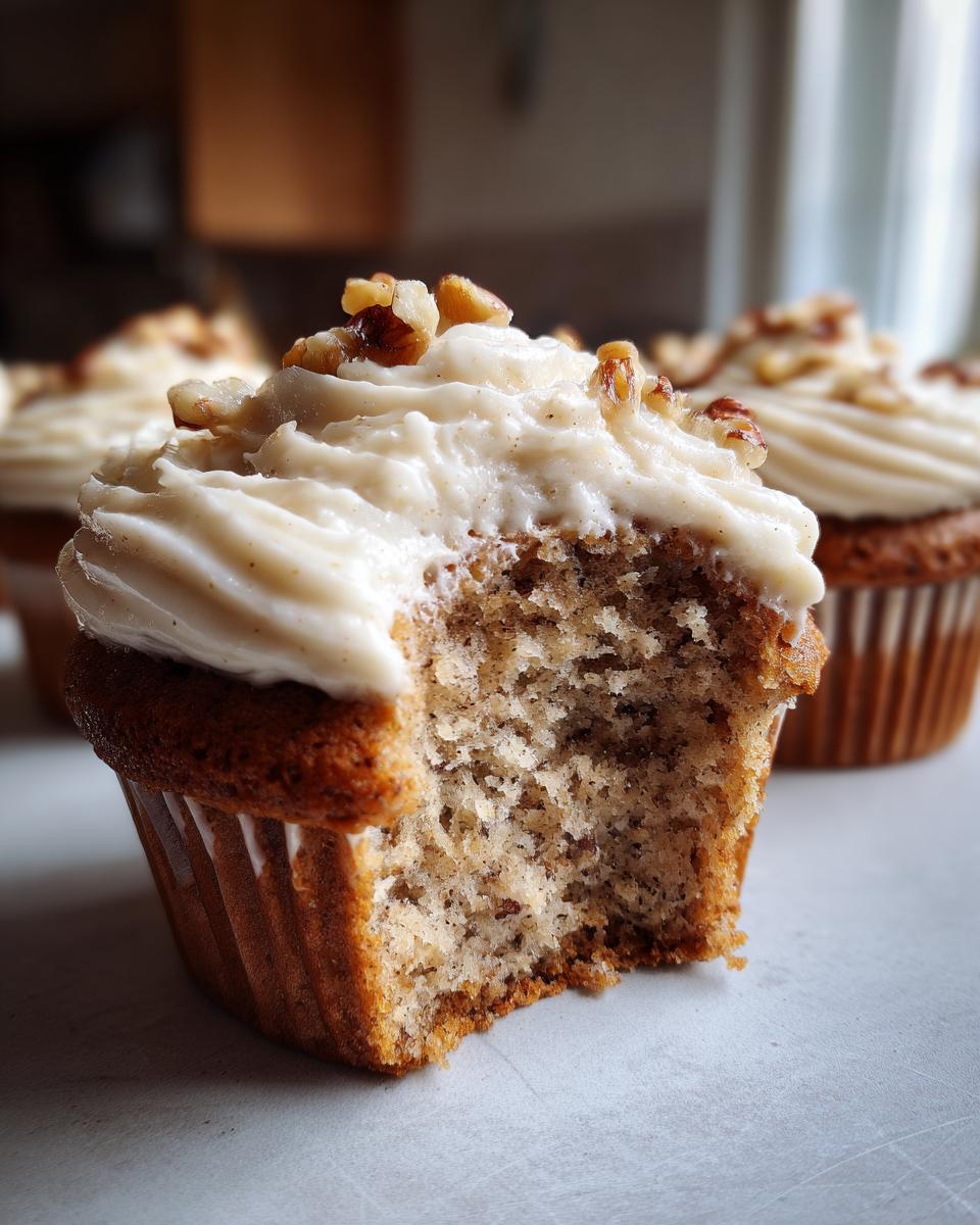 Close-up of a banana walnut cupcakes with a bite taken out, showing the moist crumb and cream cheese frosting topped with walnuts.