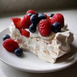 A close-up of a slice of berry pavlova showing crisp meringue, whipped cream, raspberries, and blueberries.