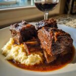 Three tender braised short ribs served over creamy polenta with rich sauce, next to a glass of red wine.