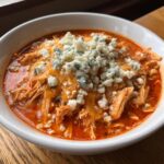 A close-up of a white bowl filled with spicy, shredded buffalo chicken chili topped generously with melted cheddar and blue cheese crumbles.