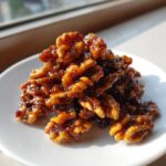 A pile of glossy, sticky candied walnuts glistening under natural light on a small white plate.