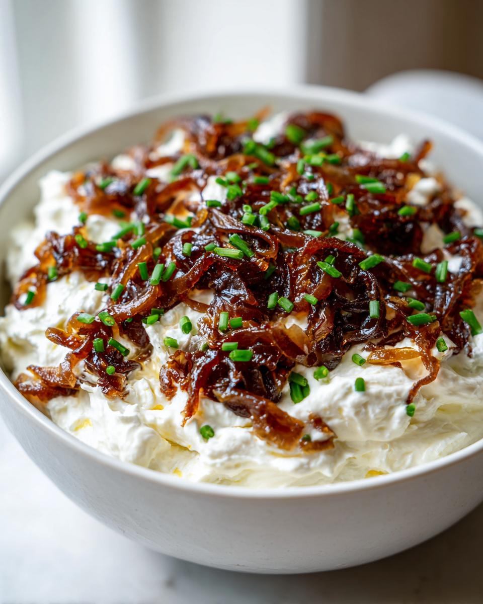 Close-up of a white bowl filled with creamy caramelized french onion dip, topped generously with dark caramelized onions and fresh chopped chives.