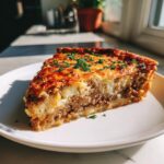 A thick slice of cheeseburger pie showing layers of ground beef and potato topping, garnished with parsley.