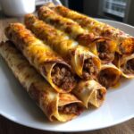 A stack of freshly baked cheesy beef flautas showing the seasoned ground beef filling and melted cheddar cheese topping.