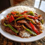 A close-up of homemade Chinese pepper steak with vibrant red and green bell peppers served over a bed of fluffy white rice.