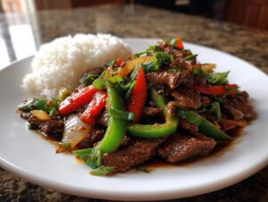 A plate of glossy Chinese pepper steak stir-fry with vibrant green and red peppers, served next to a mound of white rice.