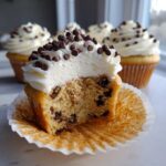 A close-up, cross-section view of a chocolate chip cookie dough cupcake showing the cake interior filled with chips and topped with white frosting and mini chips.