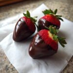 Three glossy chocolate covered strawberries resting on white parchment paper, showing bright red fruit tops.
