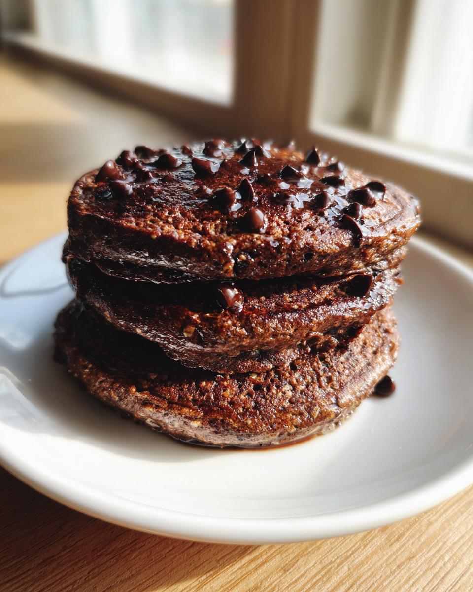 A stack of three rich, dark chocolate oat pancakes topped with melted chocolate chips and syrup.