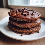 A tall stack of three rich, dark chocolate oat pancakes topped generously with mini chocolate chips.