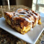 A close-up of a rich slice of cinnamon roll french toast drizzled with white icing.