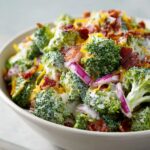 Close-up of a bowl filled with creamy apple broccoli salad, topped with bacon bits, shredded cheese, and red onion slices.