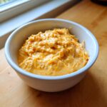 A white bowl filled with creamy, orange-colored cottage cheese queso dip, sitting on a wooden surface near a window.