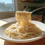 A fork lifts strands of creamy parmesan pasta from a white bowl, showing the rich sauce and grated cheese.