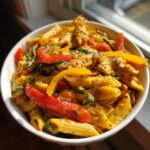 A close-up, bright photo of a white bowl filled with vibrant Rasta Pasta featuring penne, yellow and red bell peppers, and a creamy orange sauce.