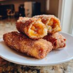 Close-up of crispy, fried peach cobbler egg rolls coated in cinnamon sugar on a white plate.