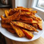 A close-up of a generous serving of perfectly seasoned, crispy sweet potato fries piled high on a white plate.