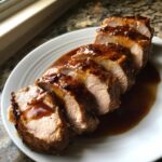 Five thick slices of juicy crockpot pork tenderloin covered in a rich, dark glaze, served on a white plate.