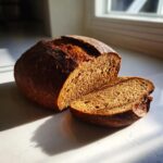 A dark, crusty loaf of dutch oven pumpernickel bread, partially sliced on a bright countertop.