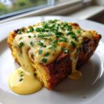 A square serving of baked eggs benedict casserole topped with creamy hollandaise sauce and fresh chives.
