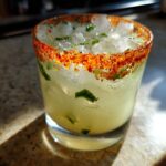 Close-up of a refreshing firecracker margarita filled with ice and jalapeño pieces, featuring a vibrant chili salt rim.