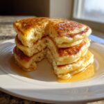 A stack of four fluffy buttermilk pancakes with a bite taken out, drizzled with syrup.