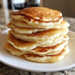 A tall stack of six fluffy buttermilk pancakes drizzled with syrup on a white plate.