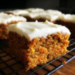 A close-up, appetizing shot of a single square of moist carrot cake bars topped with thick cream cheese frosting.