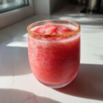 A bright pink, icy frozen firecracker rosé slushie served in a clear glass with a spiced rim.