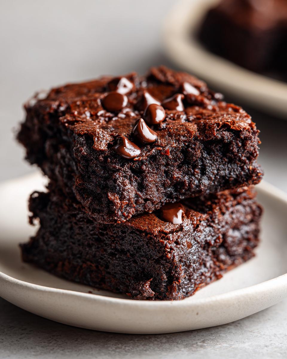 Two fudgy zucchini brownies stacked on a small white plate, topped with melted chocolate chips.