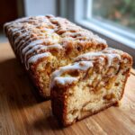 A freshly baked loaf of apple fritter bread, partially sliced, showing chunks of apple inside and drizzled with white glaze.