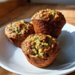 Three freshly baked zucchini muffins with visible shreds of green zucchini piled on a white plate.