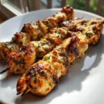 Close-up of three perfectly grilled garlic parmesan chicken skewers seasoned with herbs on a white plate.