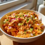 Close-up of a white bowl filled with vibrant orange healthy buffalo chicken salad, topped with diced red onion and green celery.