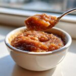 A spoonful of rich, brown spiced applesauce is lifted from a small white bowl, showing its chunky texture.
