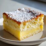 A perfect square slice of lemon custard cake, showing layers of sponge, cream, and bright yellow custard, dusted with powdered sugar.