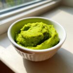 A small white bowl filled with vibrant green lemon parmesan edamame purée, sitting on a sunlit windowsill.
