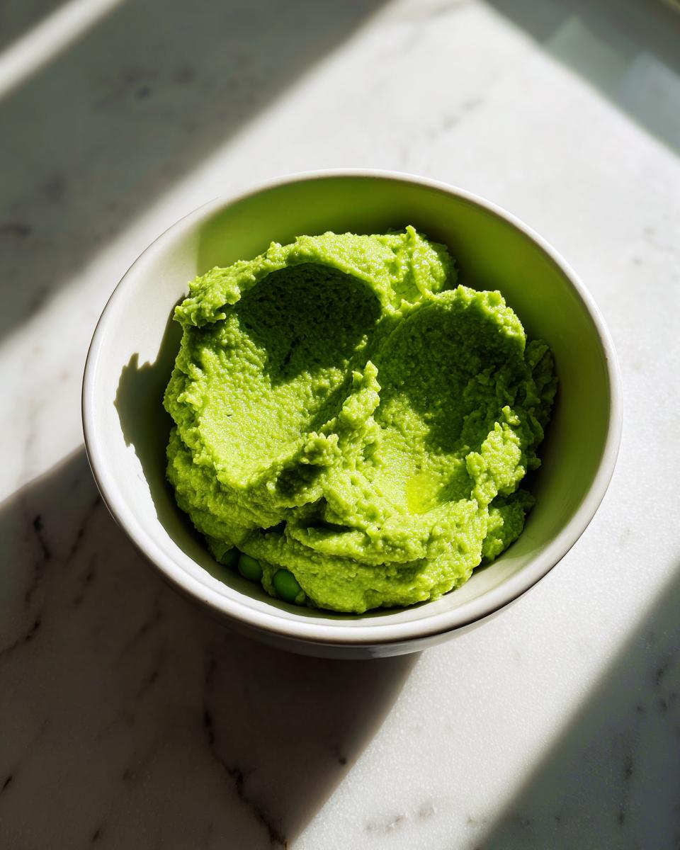 A vibrant green bowl of thick lemon parmesan edamame purée sitting on a white marble surface with strong shadows.