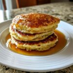 A close-up of a stack of three fluffy maple ginger pancakes soaking in golden syrup on a white plate.