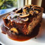 A thick slice of savory meatloaf generously covered in rich mushroom gravy and sliced mushrooms.