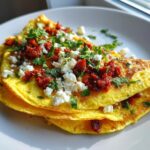Close-up of a bright yellow Mexican omelette recipe folded over, topped with crumbled white cheese, red salsa/chorizo, and fresh cilantro.
